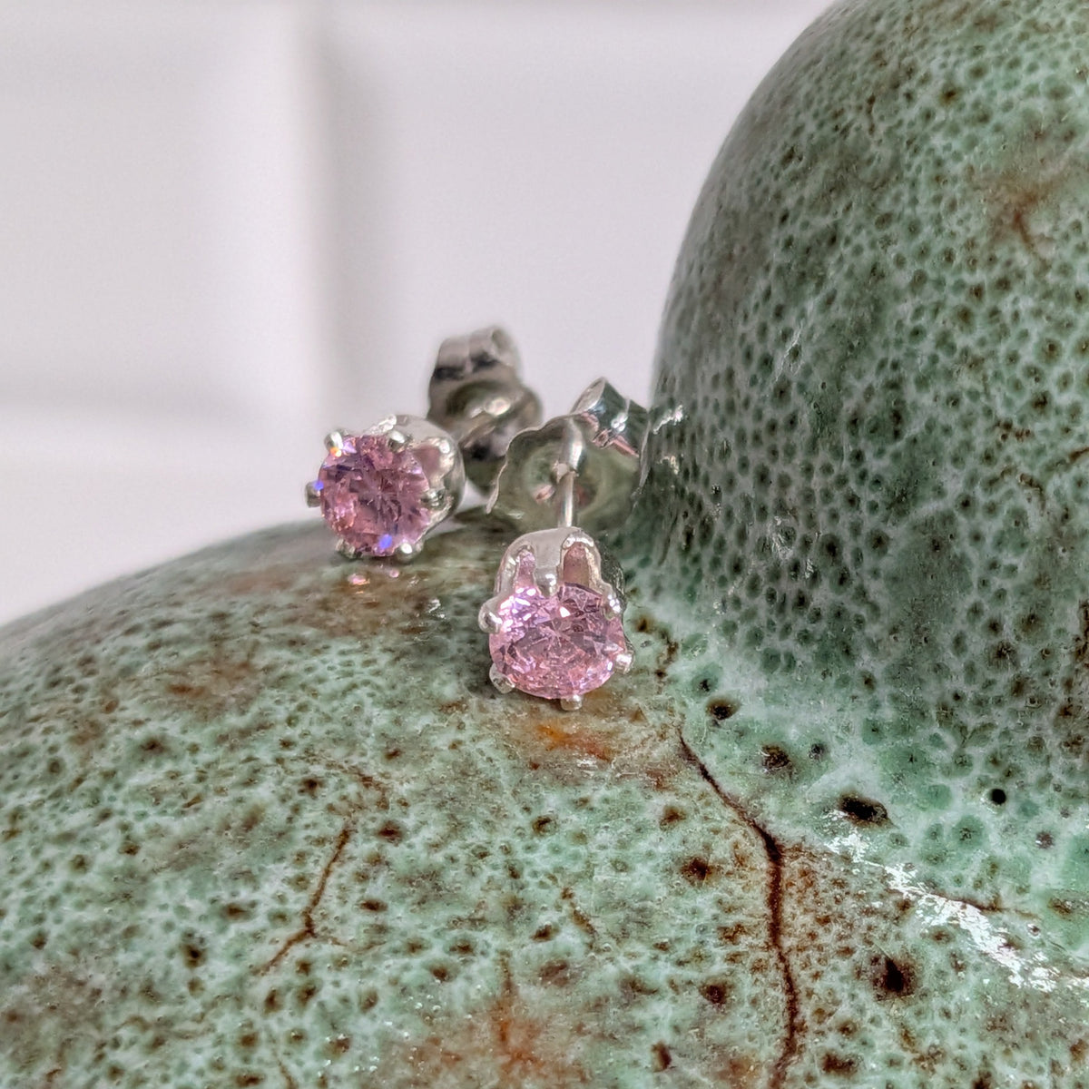 Silver stud earrings with round pink gemstones displayed on a textured green ceramic bust.