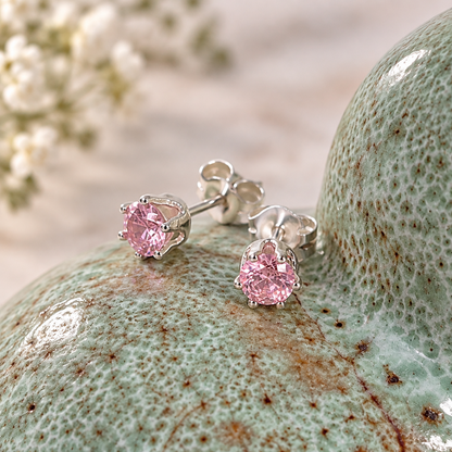 A pair of silver stud earrings with round pink gemstones, displayed on a textured light green ceramic surface.