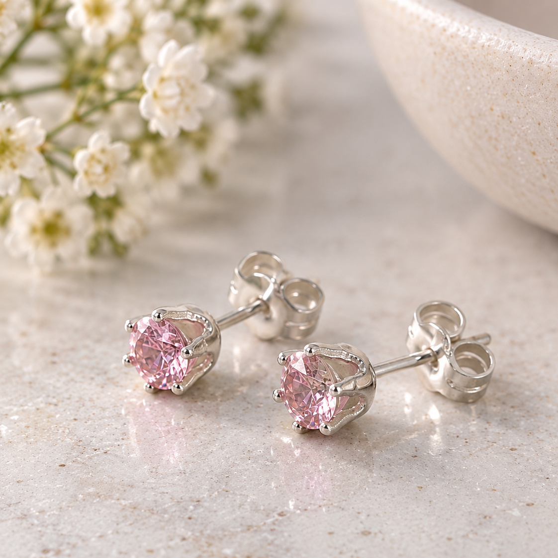 A pair of silver stud earrings with round pink gemstones, displayed on a light marble surface with small white flowers and a ceramic bowl in the background.