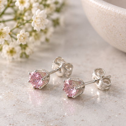 A pair of silver stud earrings with round pink gemstones, displayed on a light marble surface with small white flowers and a ceramic bowl in the background.