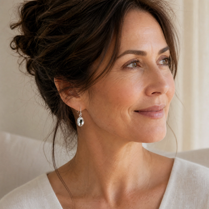 A close-up portrait of a woman with her hair in an updo, wearing a white V-neck top and a dangling silver puffed oval drop earrings set against a soft beige background.
