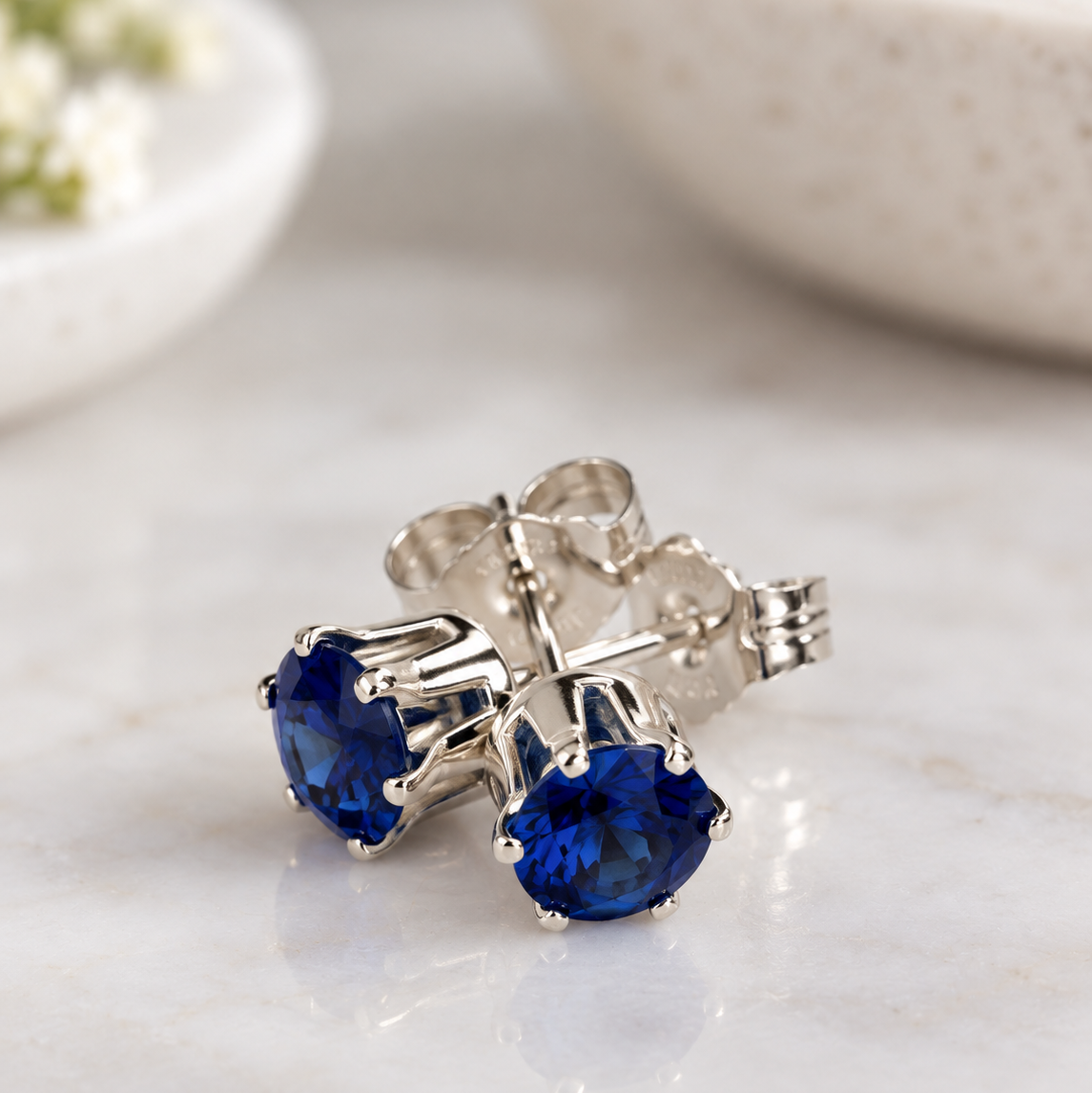 A pair of silver stud earrings with round deep blue sapphire gemstones, displayed on a light marble surface with a blurred ceramic bowl in the background.