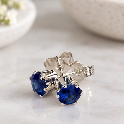 A pair of silver stud earrings with round deep blue sapphire gemstones, displayed on a light marble surface with a blurred ceramic bowl in the background.
