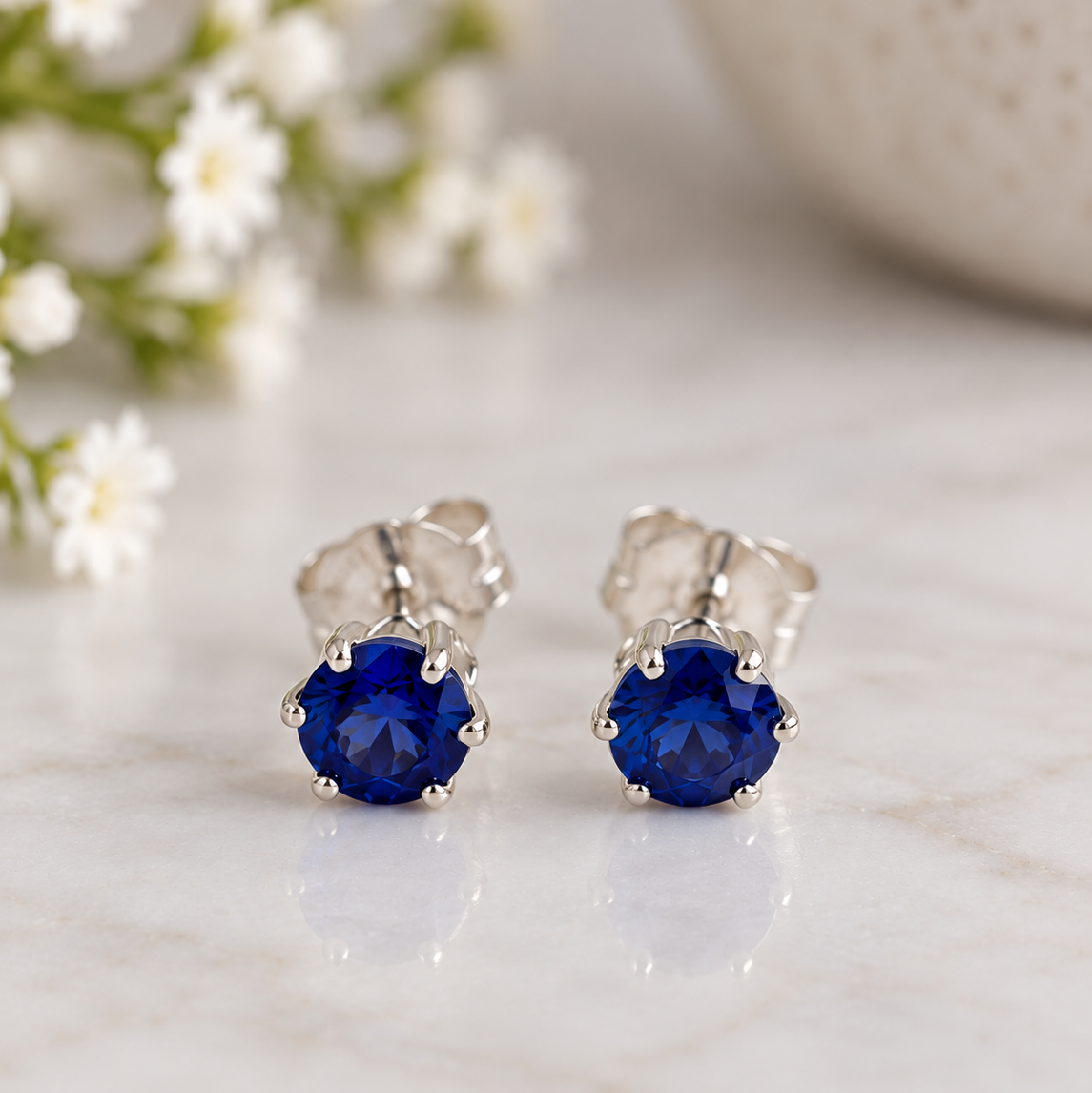 A pair of silver stud earrings with round sapphire gemstones, displayed on a light marble surface with soft-focus white flowers in the background.