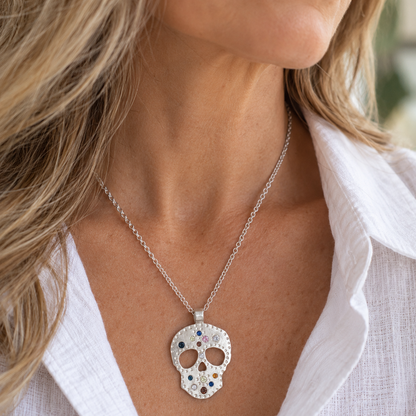 A close-up of a woman wearing a silver skull pendant necklace with small colorful gemstone accents, paired with a white linen shirt.