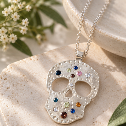 A silver skull pendant necklace adorned with colorful gemstones, resting on a textured beige stone block with small white flowers in the background.