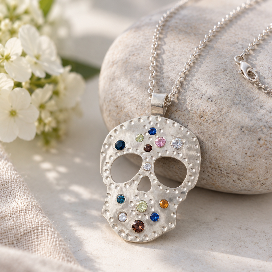 A silver sugar skull pendant necklace adorned with small multicolored gemstones, displayed on a textured stone and beige fabric with white flowers in the background.