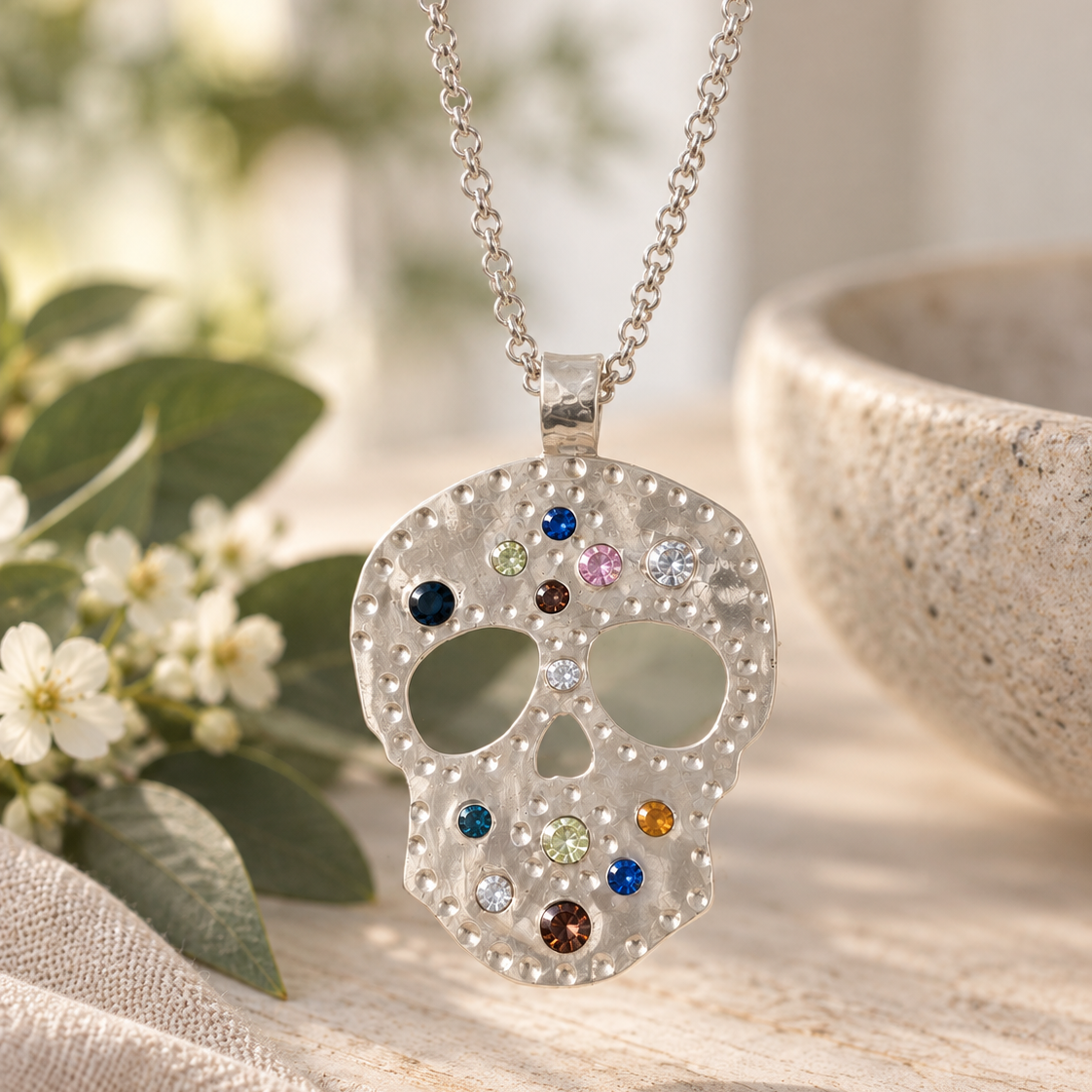 A silver skull pendant necklace adorned with small clear rhinestones and colorful gem accents, hanging on a silver chain against a softly blurred background with white flowers and a stone bowl.