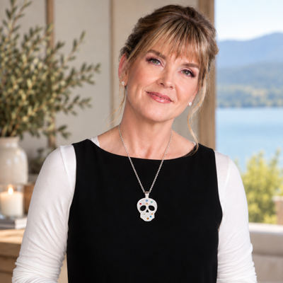 A woman wearing a black sleeveless dress over a white long-sleeve top, featuring a silver-toned necklace with a sugar skull pendant, standing in a bright indoor setting with a window view of water and mountains.