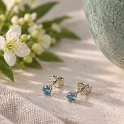 A pair of silver stud earrings with round blue gemstones resting on a textured beige fabric surface, styled with white flowers and a ceramic object in the background.