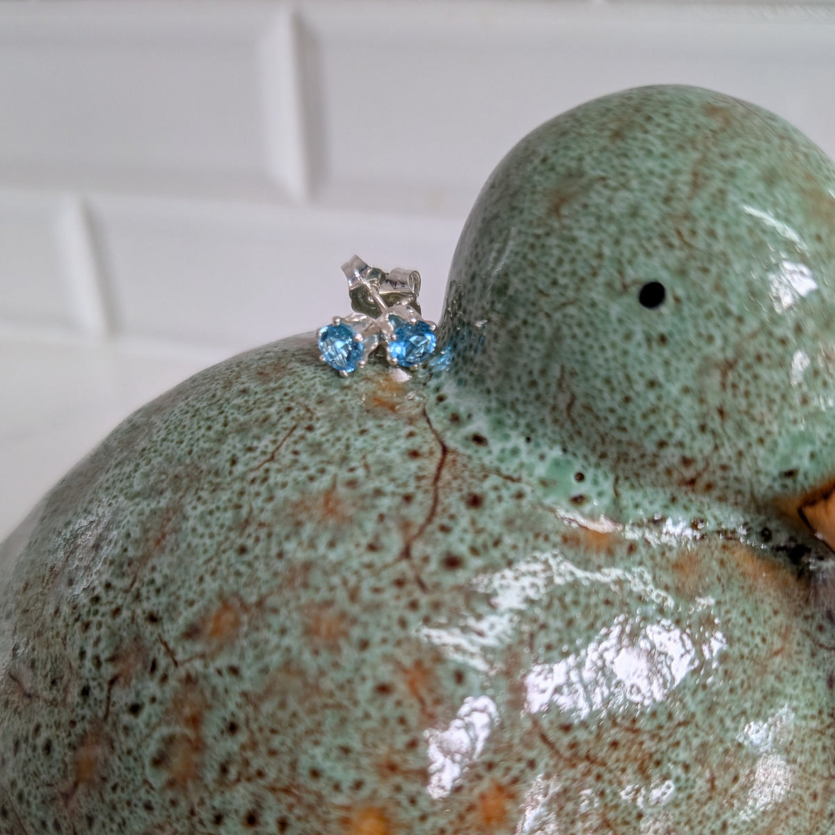 A handmade ceramic duck figurine with a glossy speckled mint green and brown glaze, featuring a small silver and blue gemstonel earring on its left shoulder, set against a white tiled background.