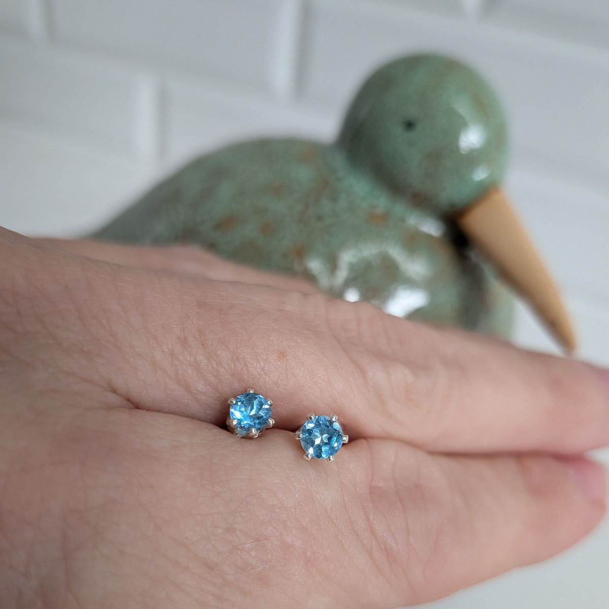A close-up of a person's hand wearing a pair of small stud earrings with light blue gemstones in silver settings, with a blurred green ceramic duck figurine in the background.