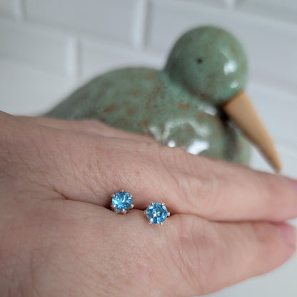 A close-up of a person's hand wearing a pair of small stud earrings with light blue gemstones in silver settings, with a blurred green ceramic duck figurine in the background.