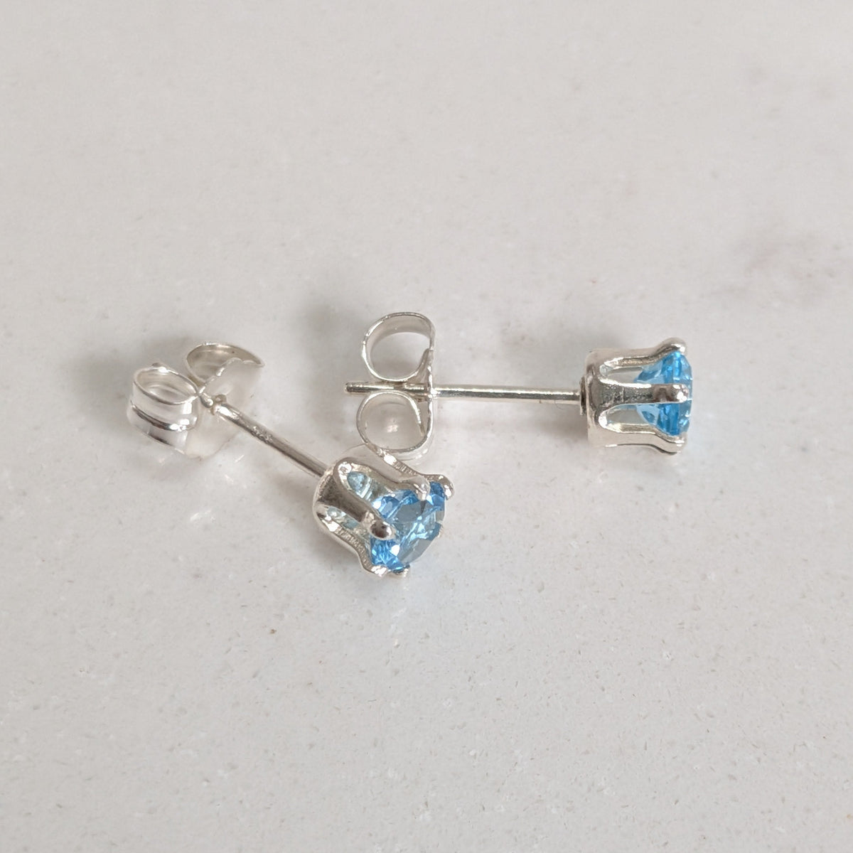 A pair of silver stud earrings with small, round-cut light blue gemstones, shown on a light gray textured surface.