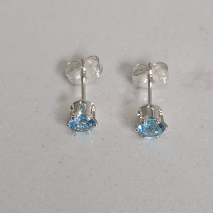 A pair of silver stud earrings with small light blue gemstones, shown on a light gray textured surface.