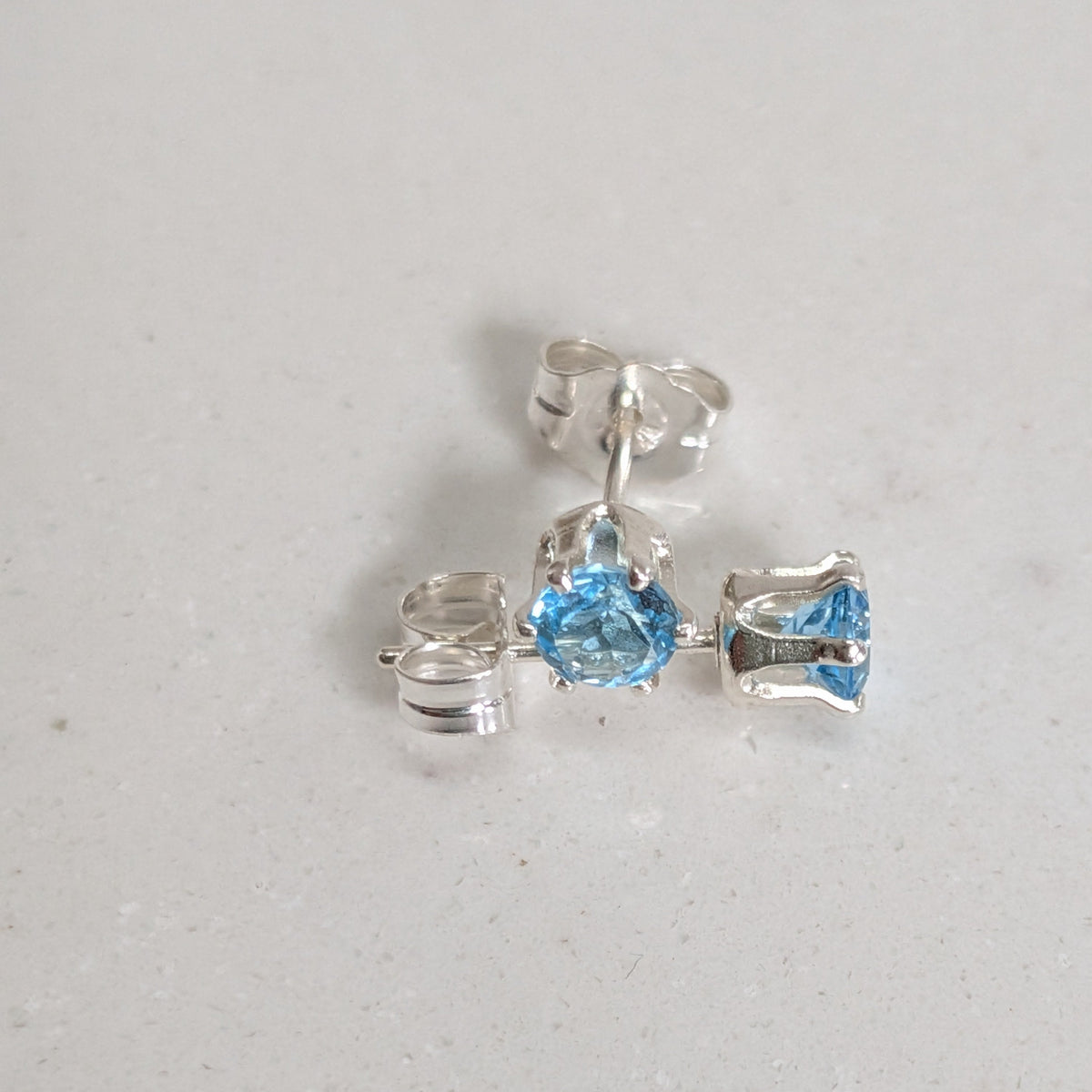 A pair of silver stud earrings with light blue gemstones, shown on a light gray textured surface. One earring is centered and fully visible, while the other lies to the left, showing its butterfly back.