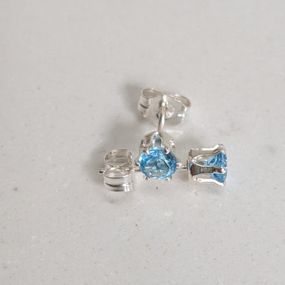 A pair of silver stud earrings with light blue gemstones, shown on a light gray textured surface. One earring is centered and fully visible, while the other lies to the left, showing its butterfly back.