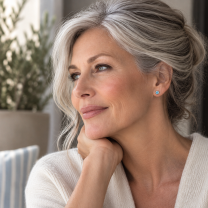 A close-up portrait of a smiling woman with gray hair wearing a white knit cardigan and a small blue stud earring, posed indoors with a softly blurred background.